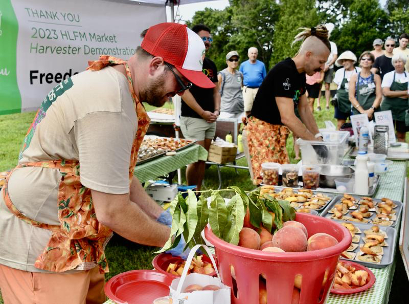 Henry Bennett of Bennett Orchards, left, slices peaches for the crowd to sample while Dru Tevis demonstrates how he makes drop-style biscuits during one of several chef workshops at the market in 2023. Tevis, SoDel Concepts corporate pastry chef, is a winner of the Food Network Holiday Baking Championship.